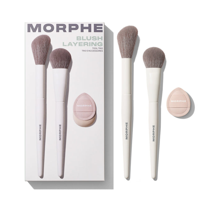 Morphe Blush Layering Tool Trio box reading "MORPHE BLUSH LAYERING TOOL TRIO TRIO D'ACCESSOIRES" beside two cream-handled taupe-bristle MORPHE brushes and a pink teardrop MORPHE powder puff.