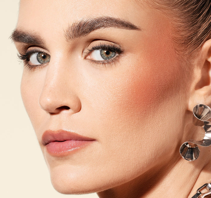 {Power Drip} Model wearing Hot Shot Blush Drops in shade Power Drip, showing a warm coral, glowing flush on cheekbones.
