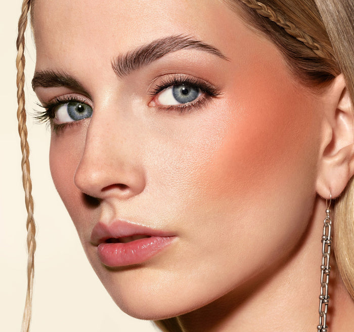 {Power Drip} Hot Shot Blush Drops in Power Drip warm peachy-coral shade swatched on cheeks of a fair-skinned model with braided hair.