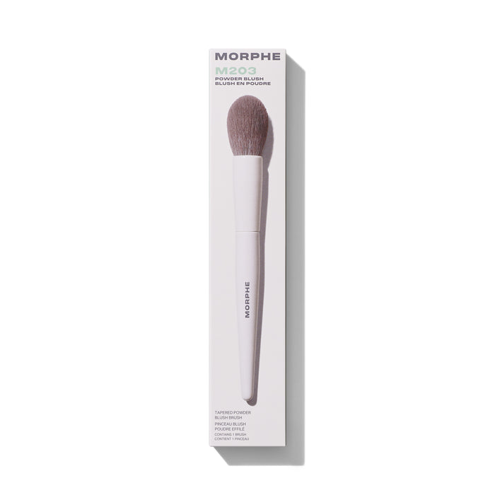 Morphe M203 Tapered Powder Blush Brush in white retail box labeled "MORPHE M203 POWDER BLUSH BLUSH EN POUDRE TAPERED POWDER BLUSH BRUSH PINCEAU BLUSH POUDRE EFFILÉ CONTAINS 1 BRUSH CONTIENT 1 PINCEAU" with mauve bristles and white MORPHE-stamped handle.